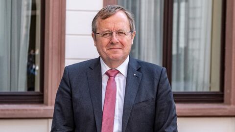 Der Hessische Raumfahrtkoordinator, Prof. Dr. Johann Dietrich Wörner, lächelt im Freien vor einem Gebäude in eine Kamera. Er trägt einen dunklen Anzug mit Hemd und roter Krawatte.